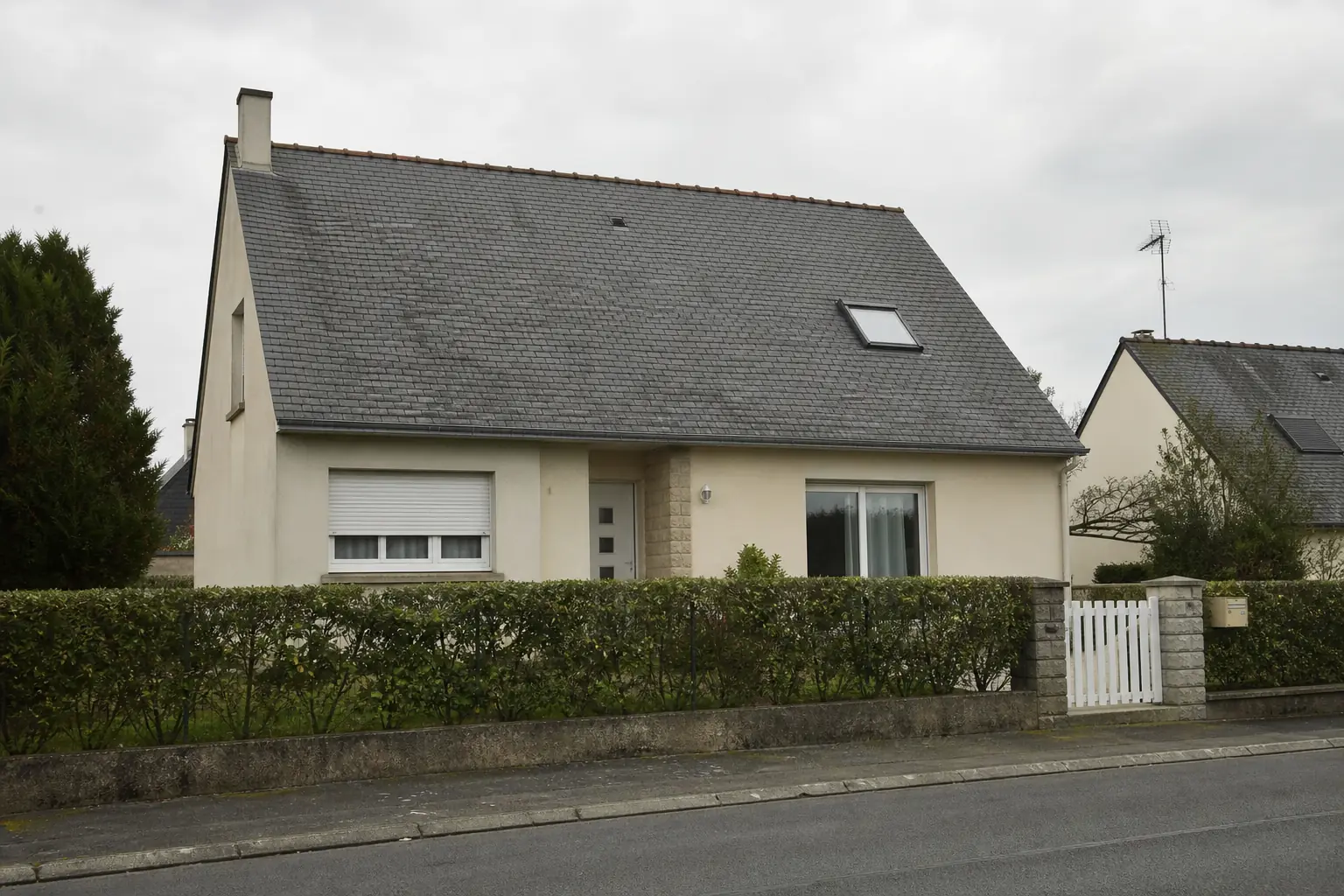 Pavillon des années 1970 vu de la rue, enduit clair, toiture d'ardoise à deux pans et haie basse