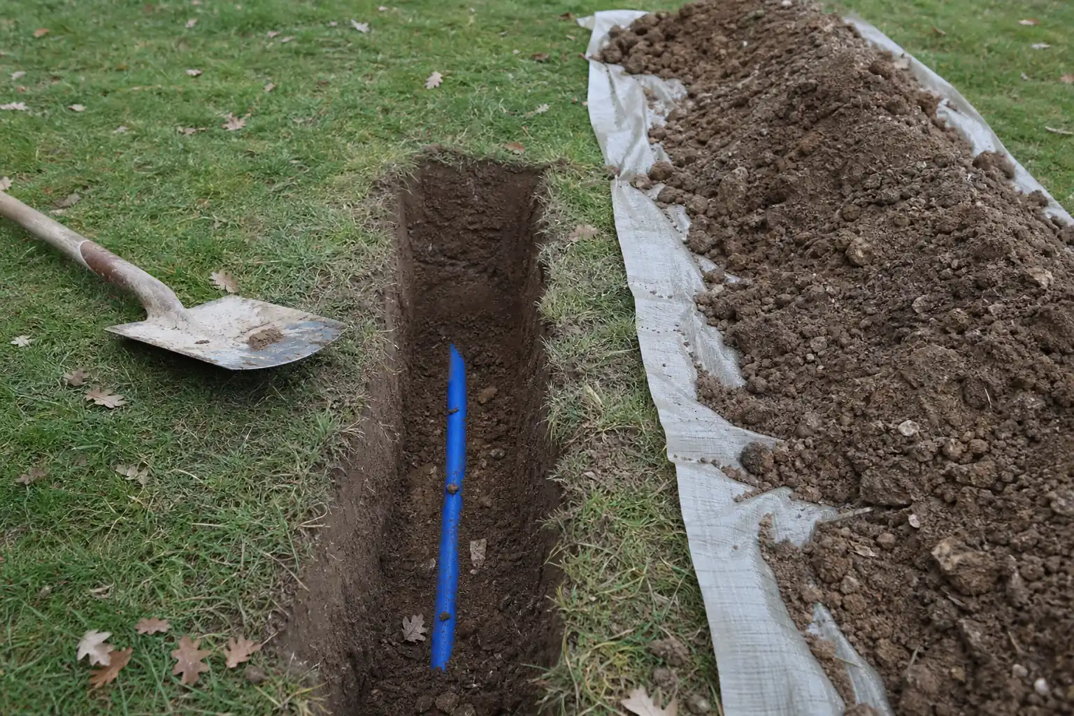 Tranchée étroite ouverte dans une pelouse, tuyau polyéthylène bleu apparent au fond, terre rangée sur une bâche